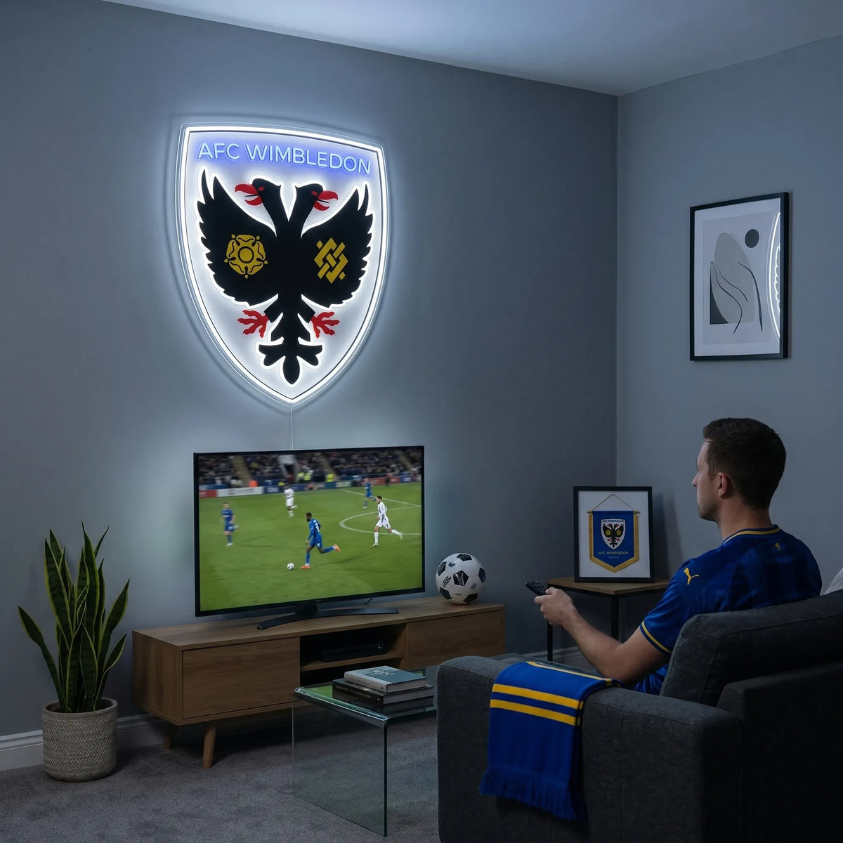 A man holding a shield-shaped AFC Wimbledon football club crest LED neon sign featuring the double-headed eagle logo glowing in a bright white and blue light