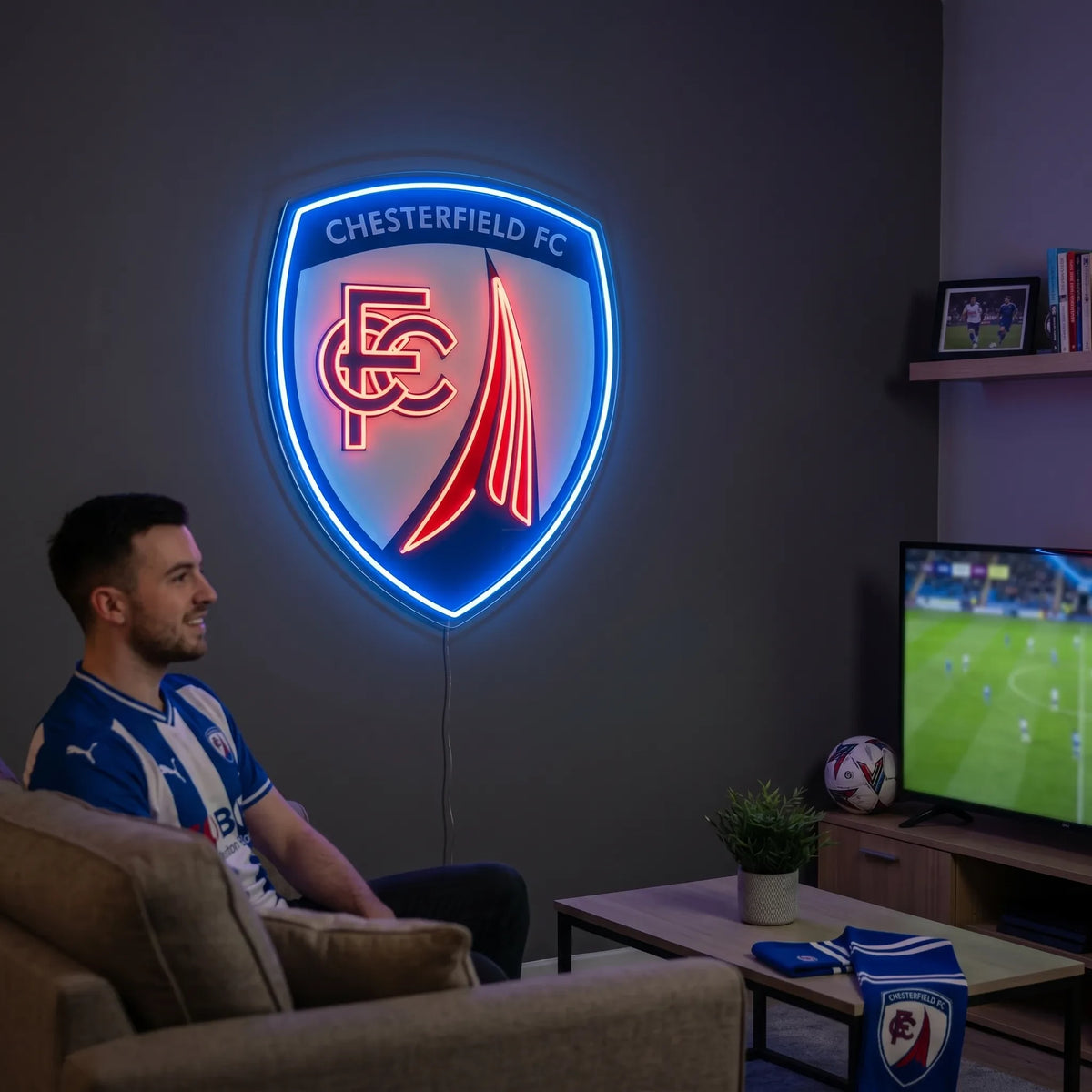 A man smiling and holding a large shield-shaped Chesterfield FC football club crest LED neon sign featuring the CFC monogram and the famous crooked spire design glowing in blue and red light