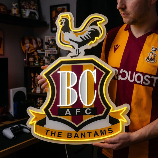 A man holding a custom Bradford City AFC football club crest LED neon sign featuring the BC initials, a bantam rooster on top, and The Bantams ribbon glowing in warm yellow and white light