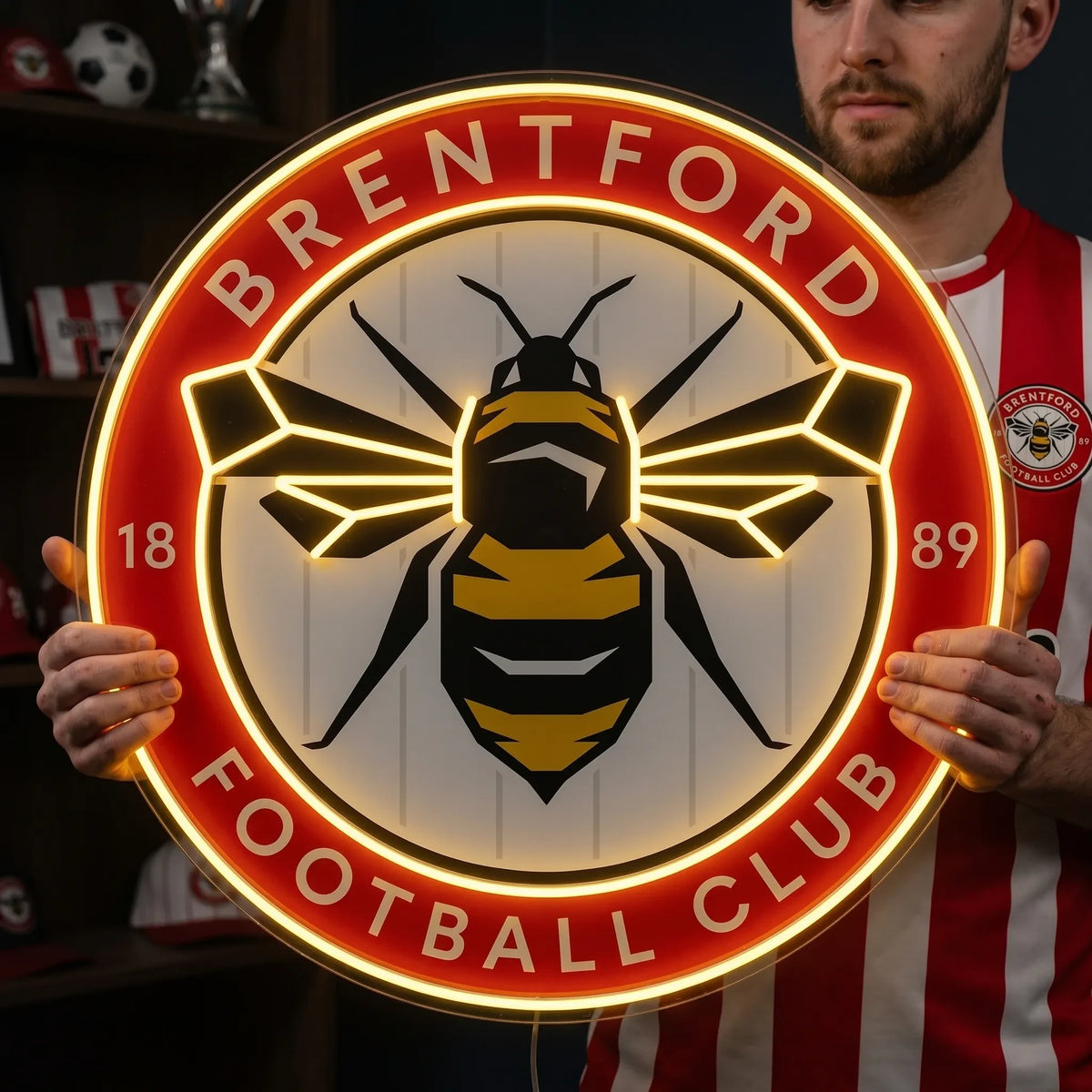 A man holding a circular Brentford FC football club crest LED neon sign featuring the iconic bee logo glowing in warm yellow and red light