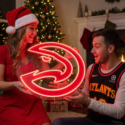 Woman in a Santa hat giving an Atlanta Hawks LED neon sign as a Christmas gift beside a decorated tree