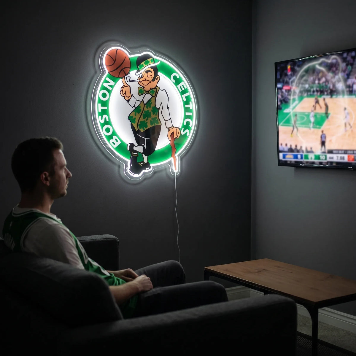 Boston Celtics LED neon sign - A fan relaxes on a couch in a dark living room beside a glowing Boston Celtics logo neon sign and a TV showing basketball.