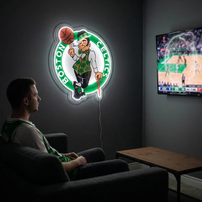 Boston Celtics LED neon sign - A fan relaxes on a couch in a dark living room beside a glowing Boston Celtics logo neon sign and a TV showing basketball.