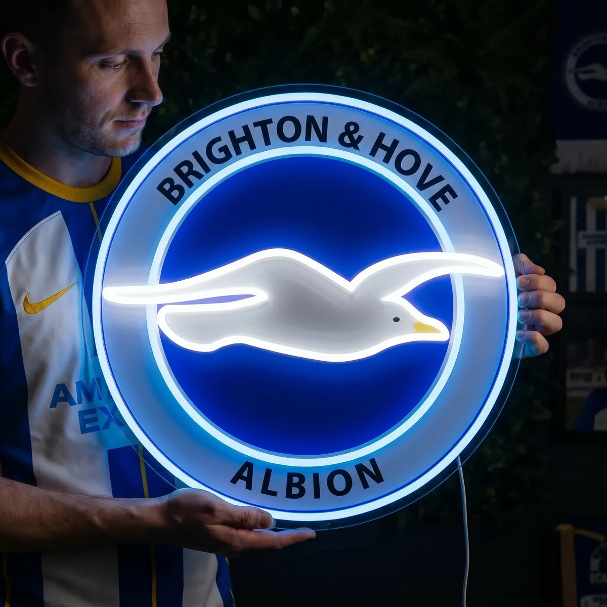 Brighton & Hove Albion FC LED neon sign - A man in a matching jersey holds a glowing blue and white round seagull crest neon sign in a fan room.