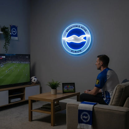 Brighton & Hove Albion FC LED neon sign - A fan relaxes on a sofa watching a football match on TV beside a glowing blue and white round seagull crest neon sign on the wall.