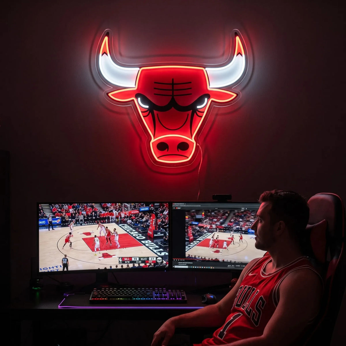 Chicago Bulls LED neon sign - A fan in a red Bulls jersey watches basketball on dual monitors under a glowing Chicago Bulls logo neon sign.