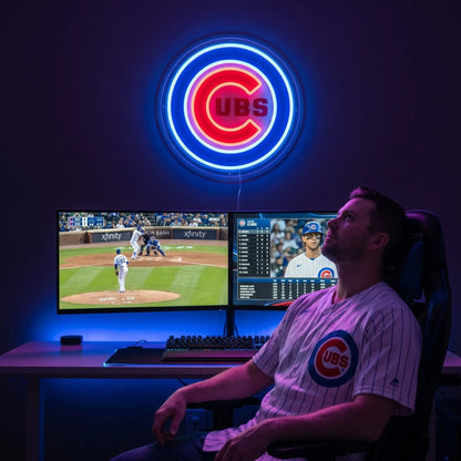 Chicago Cubs LED neon sign - A fan in a Cubs jersey watches baseball on dual monitors beneath a glowing red and blue Chicago Cubs logo neon sign.