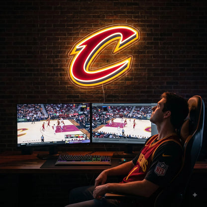 Cleveland Cavaliers LED neon sign - A fan at a gaming desk watches basketball on dual monitors below a glowing Cleveland Cavaliers C logo neon sign on a brick wall.