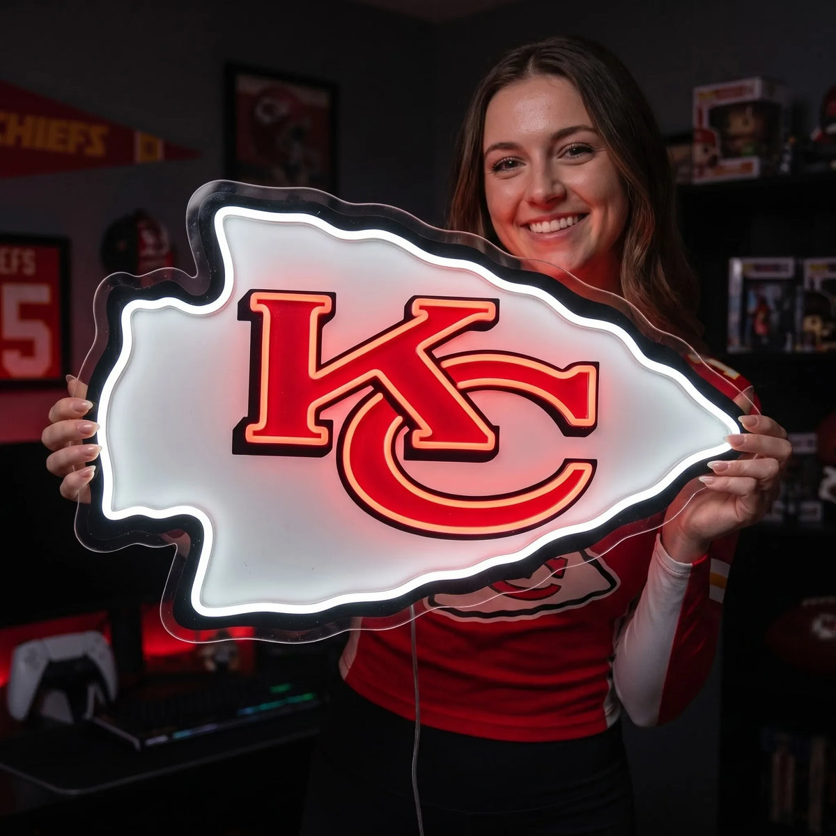 Kansas City Chiefs LED neon sign - A woman in a red Kansas City Chiefs jersey holds up a large, illuminated LED sign shaped like the team's arrowhead logo. The sign glows with a white background and features the 'KC' logo in red with black outlines.