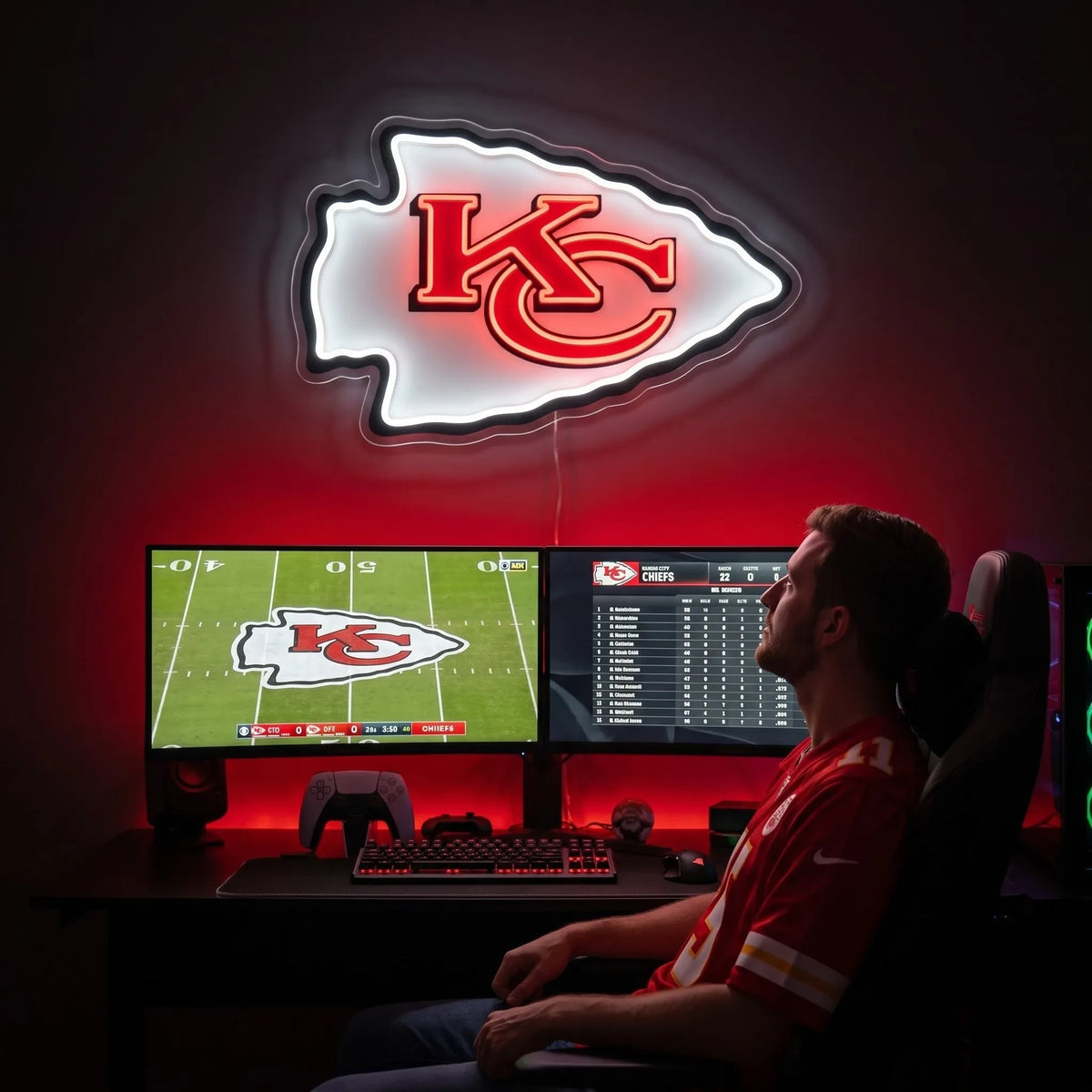 Kansas City Chiefs LED neon sign - A man sits in a gaming chair at a desk with two computer monitors, illuminated by red ambient lighting. Above the desk is a large Kansas City Chiefs NFL logo neon light glowing on a dark wall.