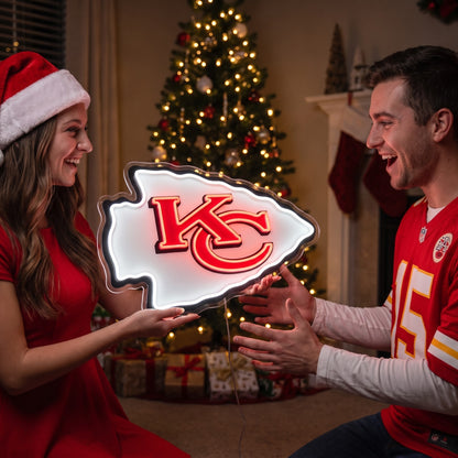 A man and a woman in holiday attire celebrate with a glowing Kansas City Chiefs arrowhead-shaped LED light in a cozy living room setting with a Christmas tree in the background.