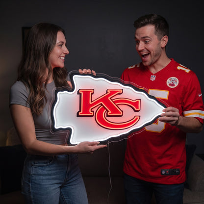 A man and a woman hold a glowing Kansas City Chiefs NFL sign shaped like the team's arrowhead logo. The man is wearing a red Kansas City Chiefs jersey, and the background is a dark, dimly lit room.