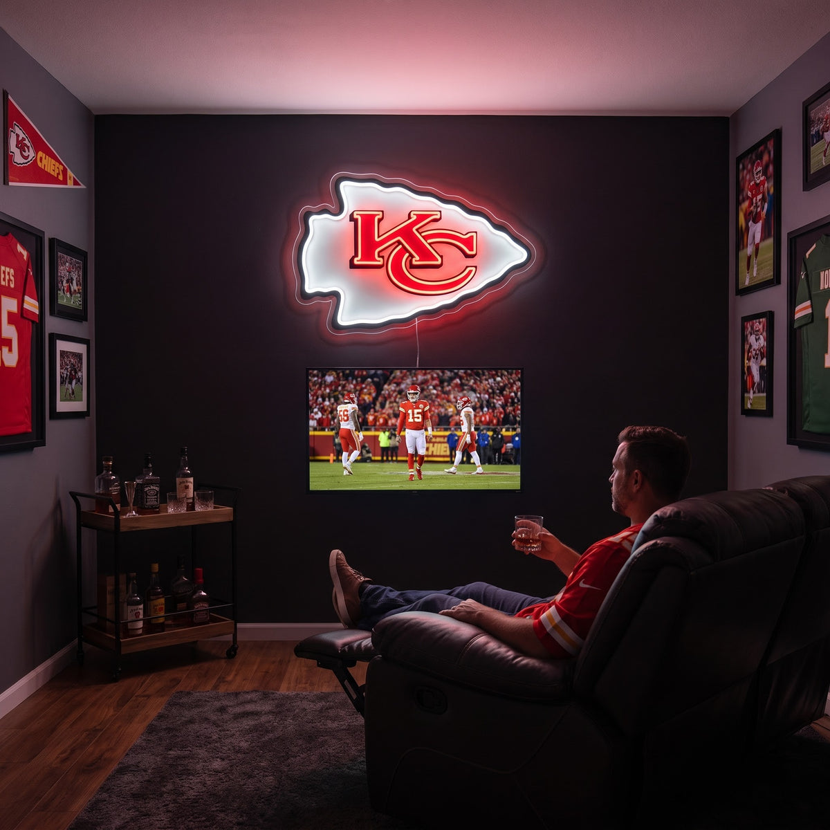 A man relaxes in a dark leather recliner watching a Kansas City Chiefs football game on TV, with a glowing Kansas City Chiefs logo mounted on the dark wall behind him.
