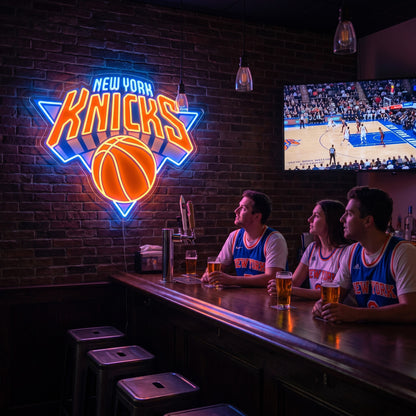 New York Knicks LED neon sign glowing orange and blue on a brick wall beside fans watching basketball at a bar