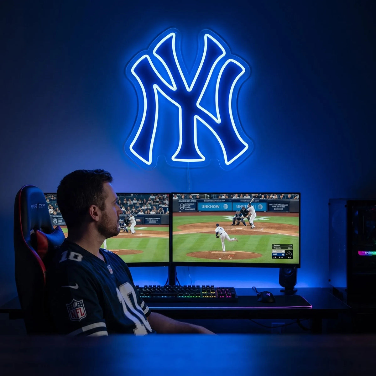 New York Yankees LED neon sign - A fan watches baseball on dual monitors under a glowing blue New York Yankees NY logo neon sign.